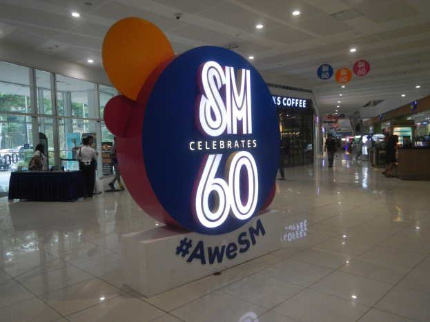 Eine Gruppe von Menschen steht auf einem Einkaufszentrumsboden unter einem "SM feiert 60-jähriges Jubiläum"-Schild, mit einem schwarzen-gedeckten Tisch links und Glastüren, Schildtafeln und Deckenleuchten im Hintergrund.