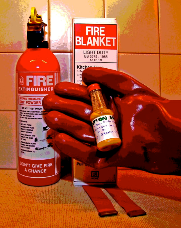 Eine rote Skulptur einer Hand mit einer Flasche rechts, ein Feuerl√∂scher links, eine Kiste in der Mitte und eine Wand im Hintergrund.