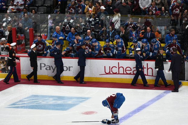 Eishockeyspieler auf dem Eis mit einem Stock, umgeben von Menschen mit Mützen, die Hockeystöcke halten, vor sitzenden und stehenden Zuschauern mit Arena-Brettern, auf denen "Colorado Avalanche vs San Jose Sharks - 3/24/15 NHL Pick, Odds, and Prediction" steht.