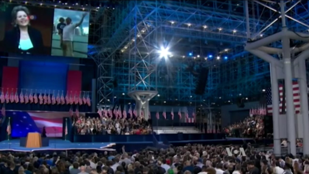 Eine große Zuschauermenge sitzt einer Bühne mit einem Rednerpult, Mikrofon, Flaggen, einem Bildschirm und einer Lichtanlage gegenüber, was auf eine Präsidentschaftsdebatte hindeutet.