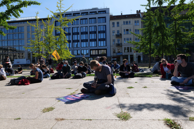 Menschen sitzen vor einem Gebäude auf dem Boden während einer Demonstration in Berlin, einige tragen Masken, mit verstreuten Taschen und Gegenständen, umgeben von Bäumen unter einem klaren blauen Himmel.