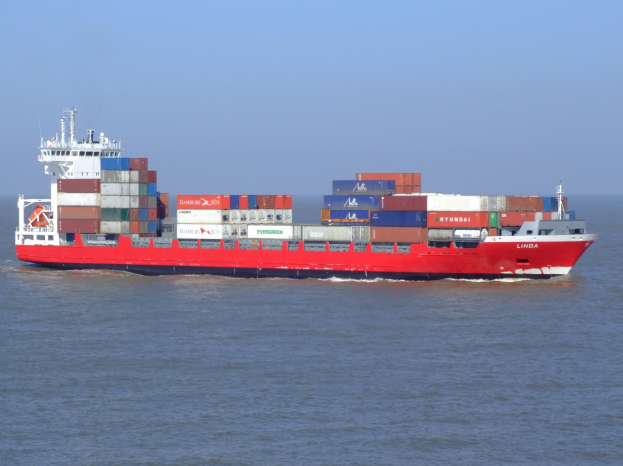 Ein großes rotes und weißes Containerschiff mit vielen gestapelten Containern segelt auf dem Meer unter einem klaren blauen Himmel.