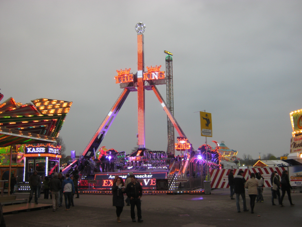 Eine Gruppe von Menschen, die nachts um eine Karussellfahrt gehen, beleuchtet von bunten Lichtern und umgeben von Buden und Bäumen, mit dem Himmel im Hintergrund.