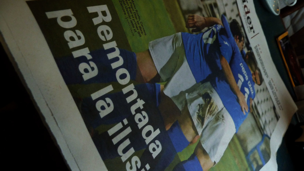 Eine Zeitung auf einem Tisch zeigt drei Männer in blauen Hemden, möglicherweise beim Fu�ballspielen, mit Text unten im Bild.