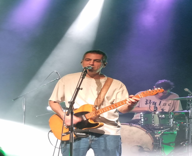 Ein Mann spielt eine E-Gitarre auf der Bühne, beleuchtet von Scheinwerfern von oben, während er in ein Mikrofon singt; ein anderer Mann sitzt hinter ihm und spielt Schlagzeug.