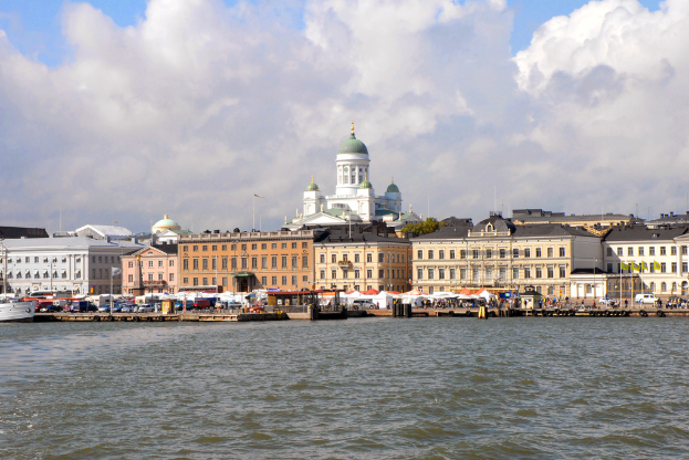 Ein braunes und weißes Gebäude mit vielen Fenstern steht vor einer Moschee mit einer grünen Kuppel, mit einem Hafen mit Booten und Frachtgut im Vordergrund und Meerwasser am Boden.