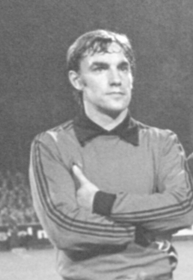 Ein Schwarz-Weiß-Foto eines älteren Mannes, der mit verschränkten Armen auf einem Fußballfeld steht und von einer Gruppe von Menschen im Hintergrund umgeben ist.