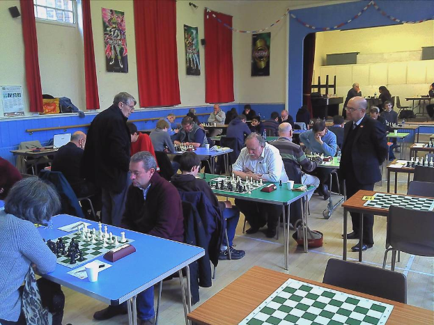 Ein großer Saal mit zwei Personen, die an einem Tisch Schach spielen, mit einem Schachbrett und Münzen darauf, Vorhängen, Postern, einem Fenster und einem Kalender an der Wand.