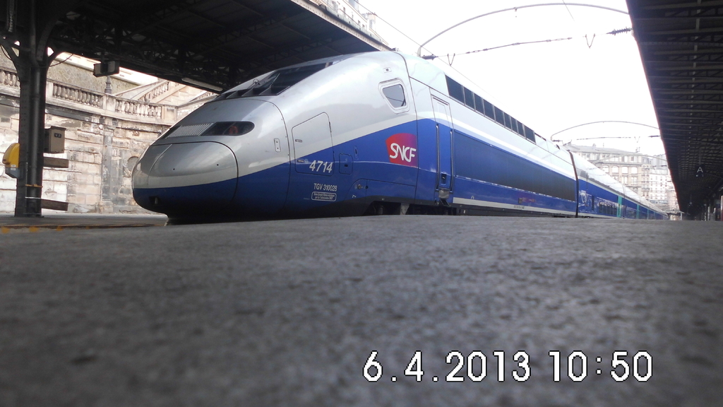 Ein Zug ist in der Mitte des Bildes zu sehen, mit einem Bahnsteig und Text unten, Gebäuden und einem Dach links und Gebäuden, Dächern, Kabeln und Himmel oben.