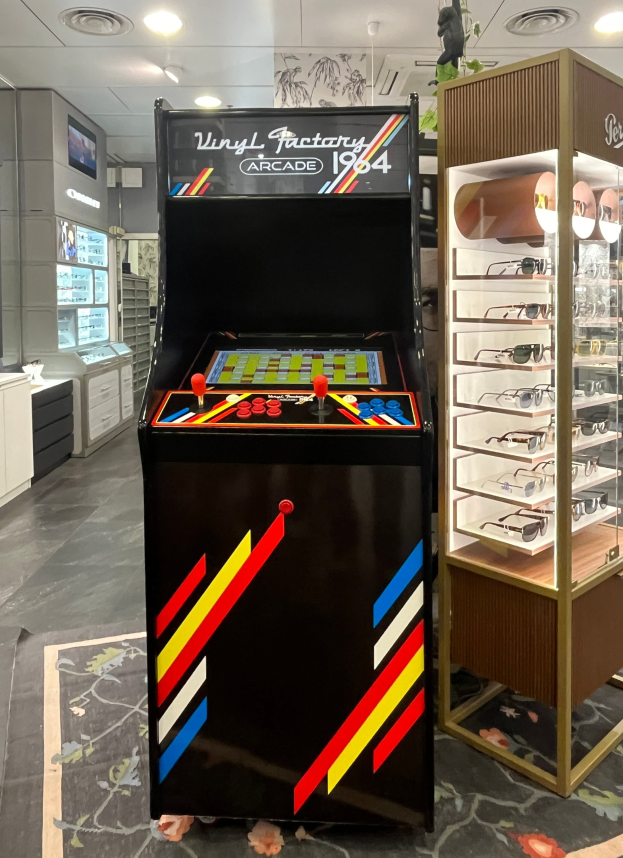 Ein Geschäft mit einem zentralen Retro-Arcade-Automaten, einem rechten Glasvitrinenschrank und Hintergrundschränken, Fernseher und Beleuchtung im Retro-Stil.