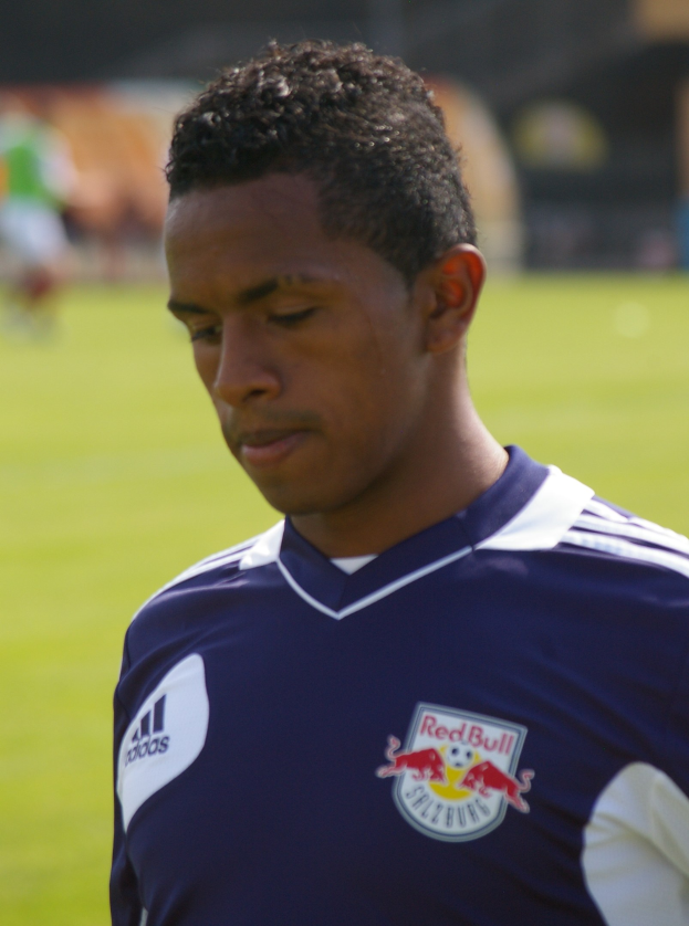 Junger Mann in blauem T-Shirt auf einem Fußballfeld, der als Flügelspieler mit kurzen dunklen Haaren und entschlossener Miene erscheint, unscharfer Hintergrund.