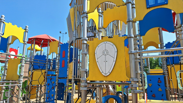 Ein Spielplatz mit buntem Spielgerät, einschließlich Stangen, Polen und einem Schirm, einige Menschen, Bäume und einen klaren blauen Himmel im Hintergrund.