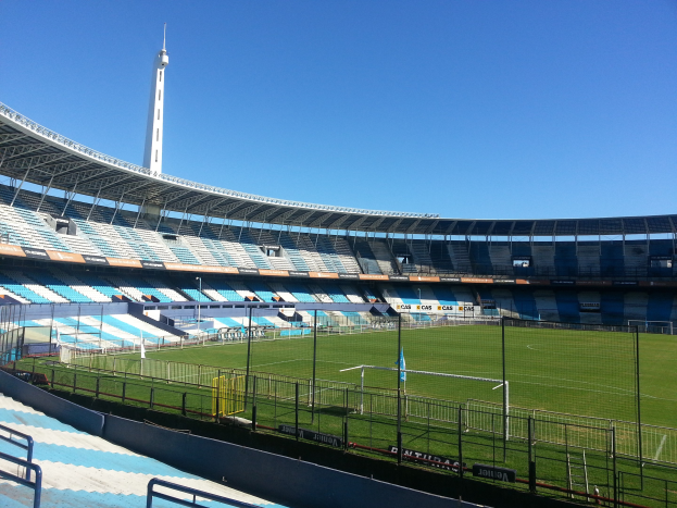 Großes Stadion mit einem von einem Zaun umgebenen Fußballfeld, einem Turm im Hintergrund und einem klaren blauen Himmel.
