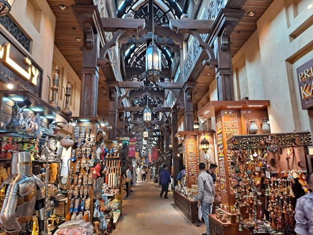 Ein belebter Dubai-Basar mit Menschen, die zwischen Regalen voller Waren hindurchgehen, beleuchtet von Deckenlampen und Wänden mit Texttafeln.