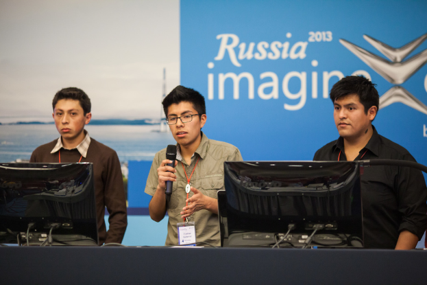 Drei Jungen stehen neben einem Schreibtisch mit zwei Computern, einer spricht in ein Mikrofon, tragen ID-Karten mit roten Tags, vor einer Wand mit einer Werbetafel, auf der "Russia Imagine 2013" steht.