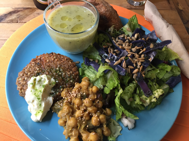 Ein blaues Teller mit einem veganen Quinoaburger und Salat, neben einem Glas Getränk, einer Flasche und einer Serviette auf einem Tisch.