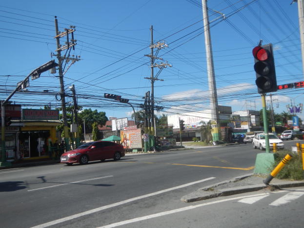 Ein rotes Auto fährt an einer Ampel vorbei auf einer Straße, die von Strommasten und -leitungen gesäumt ist, mit Gebäuden, Bäumen und einem klaren blauen Himmel im Hintergrund.
