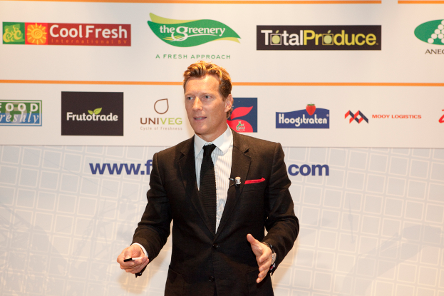 Ein Mann in einer schwarzen Blazer steht mit einem Gegenstand in der Hand und spricht, mit einem weißen Banner und einer weißen Tafel mit organisationalen Logos im Hintergrund.