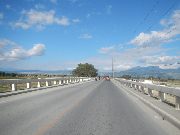 Eine Autobahn mit Motorradfahrern, umgeben von Geländern, Strommasten mit Drähten, Bäumen, Hügeln und einem bewölkten Himmel.