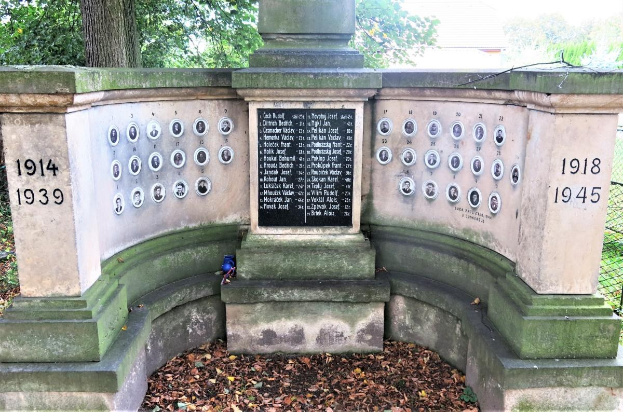 Ein Holocaust-Denkmal mit Text und Zahlen an seiner Wand, umgeben von Bäumen und einem Zaun in einem jüdischen Friedhof in Berlin, Deutschland, mit trockenen Blättern auf dem Boden.