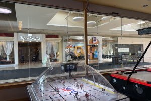 Eine Eishalle in einem Einkaufszentrum, umgeben von Glaswänden mit Menschen, Möbeln und Beschriftungen, die durch die Glaswände sichtbar sind, und einem Air-Hockey-Tisch auf der rechten Seite.