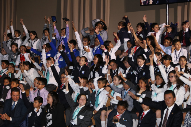 Gruppe von Kindern auf einer Bühne sitzend mit einem Mann und einer Frau davor; Bühnenhintergrund enthält Vorhänge, eine Wand und einen Fernseher auf einem Ständer.