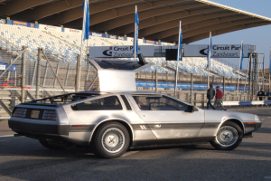 Ein silberner DeLorean DMC-12 fährt auf einer Straße, umgeben von Menschen, Fahnen, Schildern, Geländern, Stühlen und einem Schuppen, mit Bäumen und einem klaren blauen Himmel im Hintergrund.
