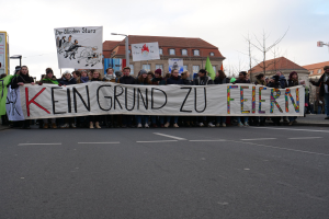 Protestierende mit einem Banner "Kein Grund zu Feiern" gegen deutsche Sparmaßnahmen, mit Gebäuden, Bäumen und einem klaren Himmel im Hintergrund.