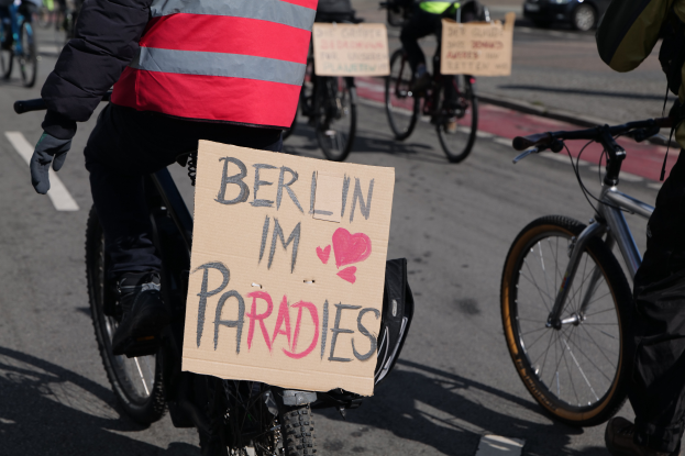 Eine Gruppe von Menschen auf Fahrrädern auf einer Straße mit einem Schild "Berlin I'm Paradies" im Vordergrund, ein Auto im Hintergrund und das Bild ist leicht unscharf.