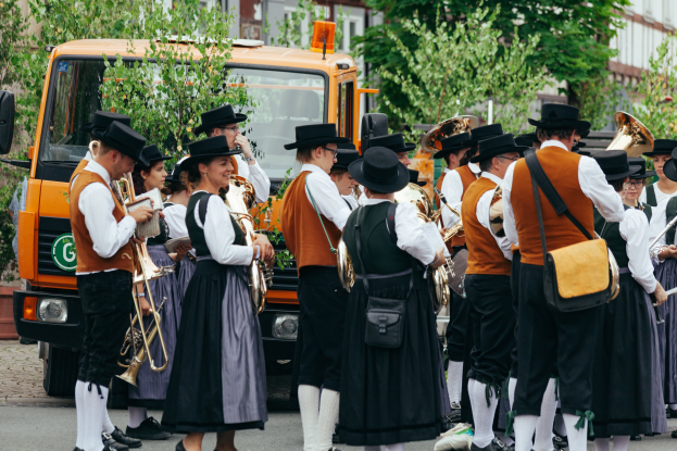 Eine Gruppe von Menschen in traditioneller bayrischer Tracht marschiert durch eine Straße, spielt Musikinstrumente und trägt Taschen, mit Bäumen, Gebäuden und einem geparkten Fahrzeug im Hintergrund.