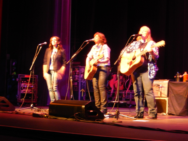 Drei Frauen performen auf der Bühne eines Musikkonzerts, jede singt in ein Mikrofon, während sie eine Gitarre spielt, mit einem Tisch voller Wasserflaschen und Gitarren dahinter.