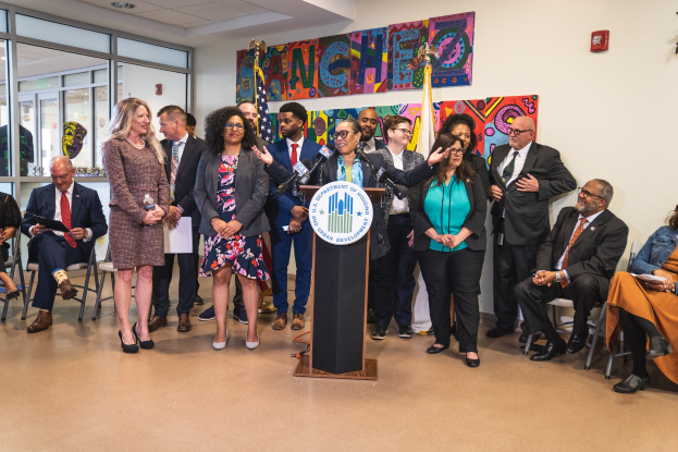 Gruppe von Menschen vor einem Podium mit einem Mikrofon und einer Tafel mit der Aufschrift "Sancho Stadtrat tritt dem Stadtrat bei", mit sitzenden und stehenden Teilnehmern, Flaggen, einem Feuerlöscher und Deckenleuchten im Hintergrund.