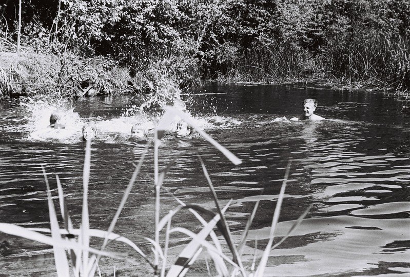 Schwarz-weiß-Foto von Menschen, die in einem Fluss umgeben von Bäumen und Pflanzen schwimmen.