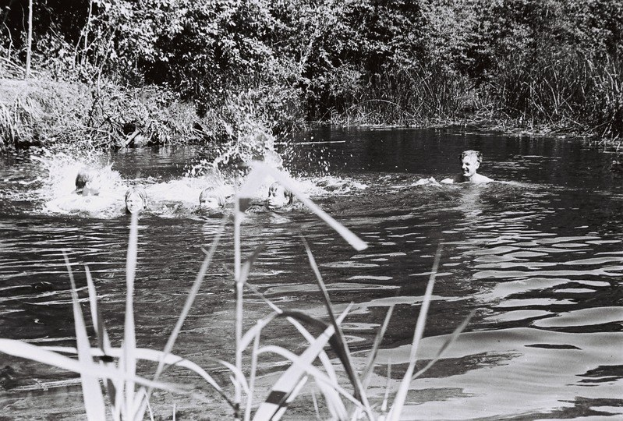 Schwarz-weiß-Foto von Menschen, die in einem Fluss umgeben von Bäumen und Pflanzen schwimmen.
