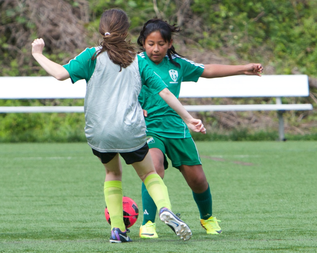 Zwei Mädchen in grünen Trikots beim Fußballspielen auf einem grünen Rasen mit einer weißen Bank, Bäumen und Pflanzen im Hintergrund und einer Wand unten.
