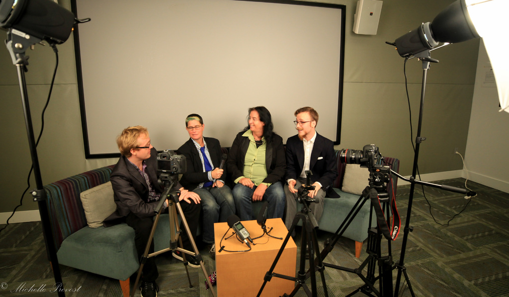 Eine Gruppe von drei Personen sitzt auf einem Sofa und gibt ein Interview. Links sitzt eine Person in schwarzer Jacke und Hose, in der Mitte eine Person in einem Mantel und einer blauen Krawatte mit einem Mikrofon und rechts eine strahlende Person. Auf dem Tisch in der Mitte steht ein Mikrofonrecorder, Kameras sind auf der rechten und linken Seite und große Scheinwerfer sind vorhanden. Im Hintergrund ist ein Projektionsschirm zu sehen.