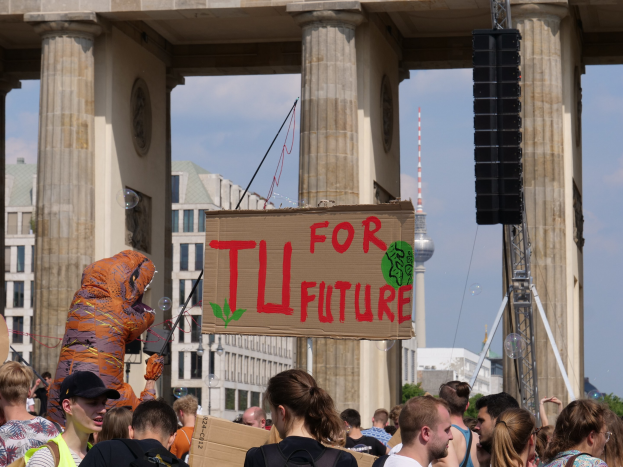 Eine Gruppe von Menschen vor dem Brandenburger Tor in Berlin, Deutschland, mit einigen, die Taschen tragen und einer im Kostüm mit einem Schild, vor einem Hintergrund von Gebäuden, Bäumen und einem Turm unter einem bewölkten Himmel.