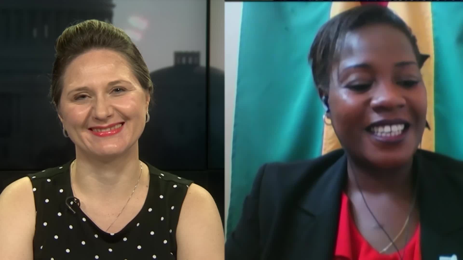 Zwei Frauen, die während eines Videoanrufs lächeln, eine in einem schwarzen und weißen Kleid, die andere in einer unbekannten Farbe, mit einer Flagge an der Wand hinter ihnen in einer öffentlichen Einrichtung.