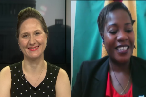 Zwei Frauen, die während eines Videoanrufs lächeln, eine in einem schwarzen und weißen Kleid, die andere in einer unbekannten Farbe, mit einer Flagge an der Wand hinter ihnen in einer öffentlichen Einrichtung.