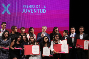 Eine Gruppe von Menschen auf einer Bühne, die strahlen und Zertifikate halten, mit einer Person in der Mitte, die ein Mikrofon hält, und einem Bildschirm im Hintergrund mit der Aufschrift "Premio de la Juventud 2017".