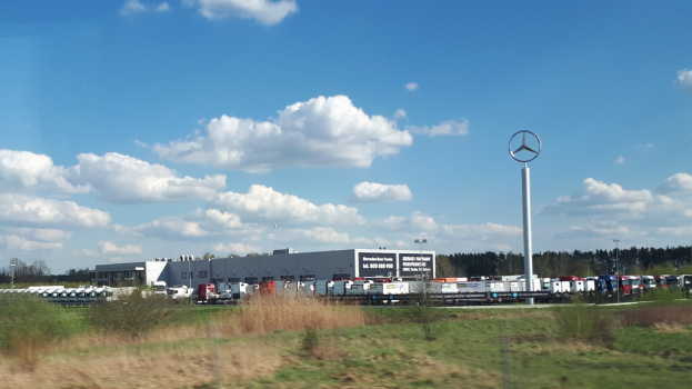 Außenansicht einer Mercedes-Benz-Fabrik in Deutschland mit Fahrzeugen auf der Straße, Grünflächen, Gebäuden und einem Logo-Pfahl unter einem bewölkten Himmel.
