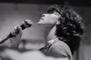 Eine Frau in einem weißen Kleid singt in ein Mikrofon auf der Bühne, ihre Augen geschlossen und ihr Gesichtsausdruck voller Emotion.