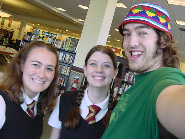 Ein Mann und zwei Studenten machen ein Selfie in einer Bibliothek, im Hintergrund sind Bücherregale mit vielen Büchern zu sehen.
