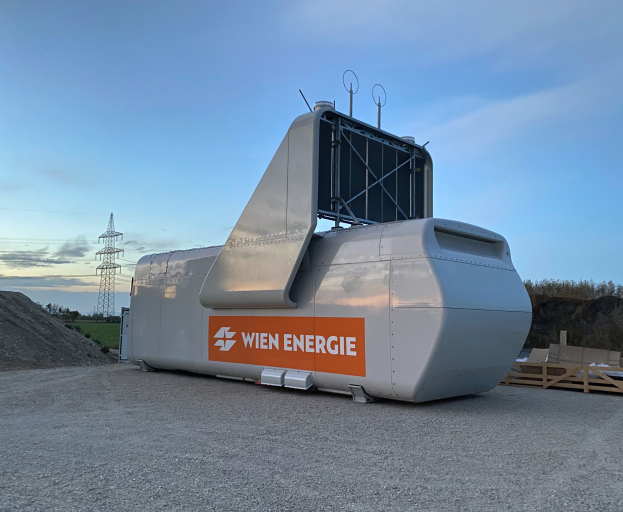Eine große weiße und orangefarbene Windturbine steht auf einem Schmutzfeld umgeben von Bäumen und einem Turm im Hintergrund, mit ein paar Gegenständen auf der rechten Seite und dem Himmel oben sichtbar.