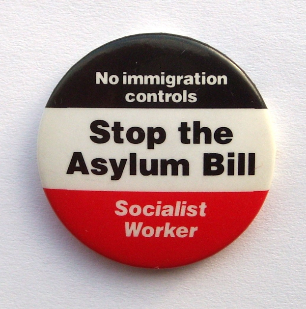Ein rundes schwarzes Abzeichen mit weißer fettgedruckter Schrift, die 'Stoppt das Asylgesetz Sozialistische Arbeiter' auf einer weißen Oberfläche liest.