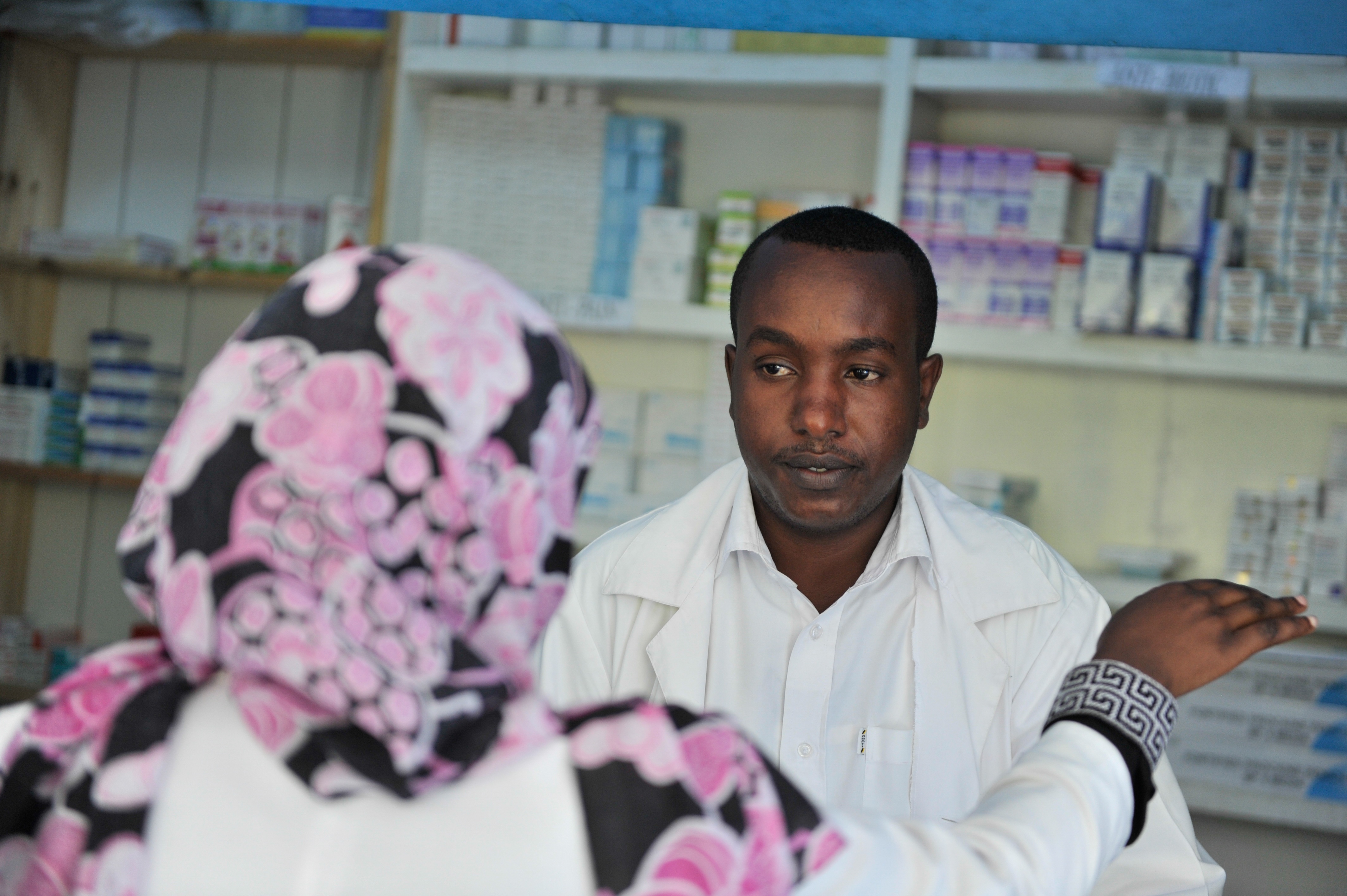 Eine Apothekerin in einem weißen Laborkittel spricht mit einer sitzenden Frau in einer Apotheke, mit Regalen voller Schachteln und anderen Gegenständen im Hintergrund.