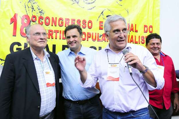 Ein Mann spricht in ein Mikrofon vor einem gelben Vorhang mit der Aufschrift "18. Grad-Kongress o", während andere Männer im Hintergrund stehen.