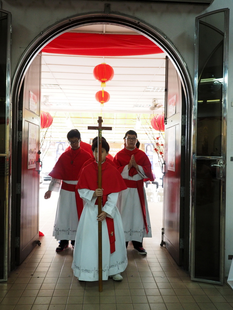 Menschen beim Gehen mit einem Kreuz in einer Kathedrale, mit Türen links und rechts und Blumen und dekorativen Elementen im Hintergrund.
