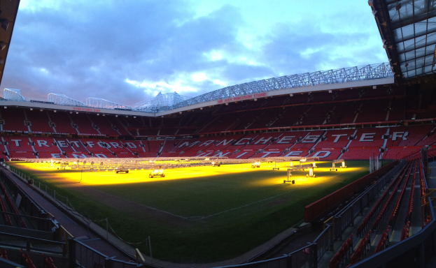 Old Trafford Stadion, Heimat von Manchester United, mit einem grünen Feld in der Mitte, umgeben von Sitzreihen, hellen Lichtern, einer hüttenähnlichen Struktur auf der rechten Seite und einem Himmel mit weißen Wolken.