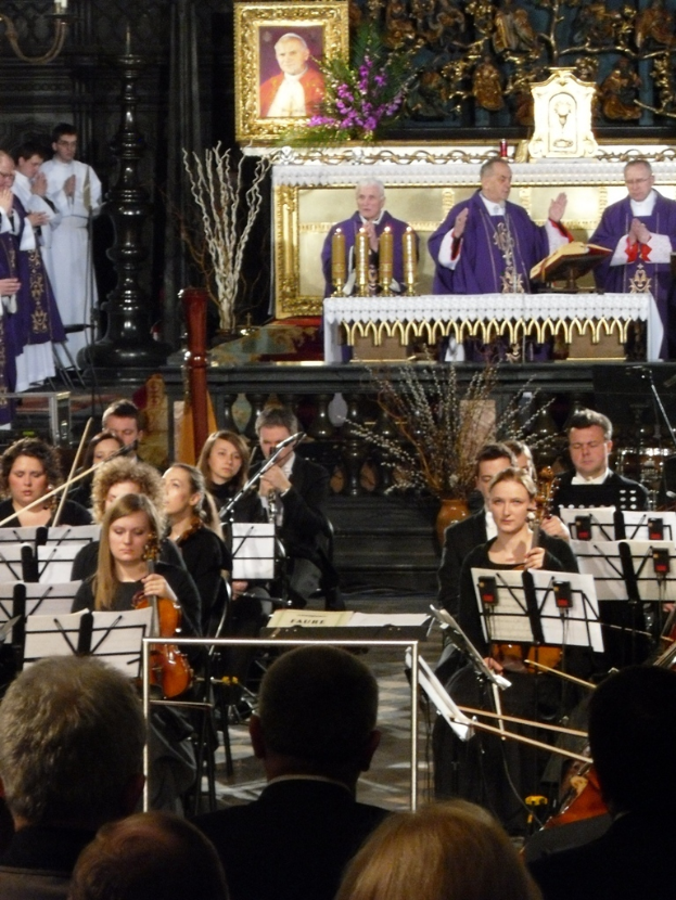 Eine Gruppe von Menschen, die Geigen spielen, mit Noten vor sich, während im Hintergrund drei Kirchenväter hinter einer Bibel stehen, mit einem von Schnitzereien umgebenen Fotorahmen dahinter.